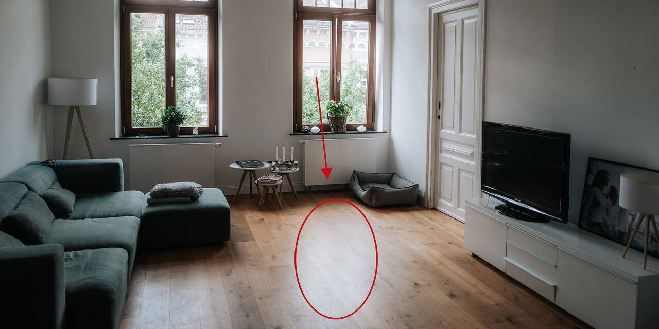Auf dem Foto ist ein Wohnzimmer mit zwei großen Fenstern zu sehen. Auf der linken Seite steht eine grüne Couch und dahinter eine Lampe. Auf der rechten Seite befindet sich ein Sideboard mit einem Fernseher und dahinter eine Tür. In der Mitte des Fotos ist ein roter Kreis eingezeichnet.