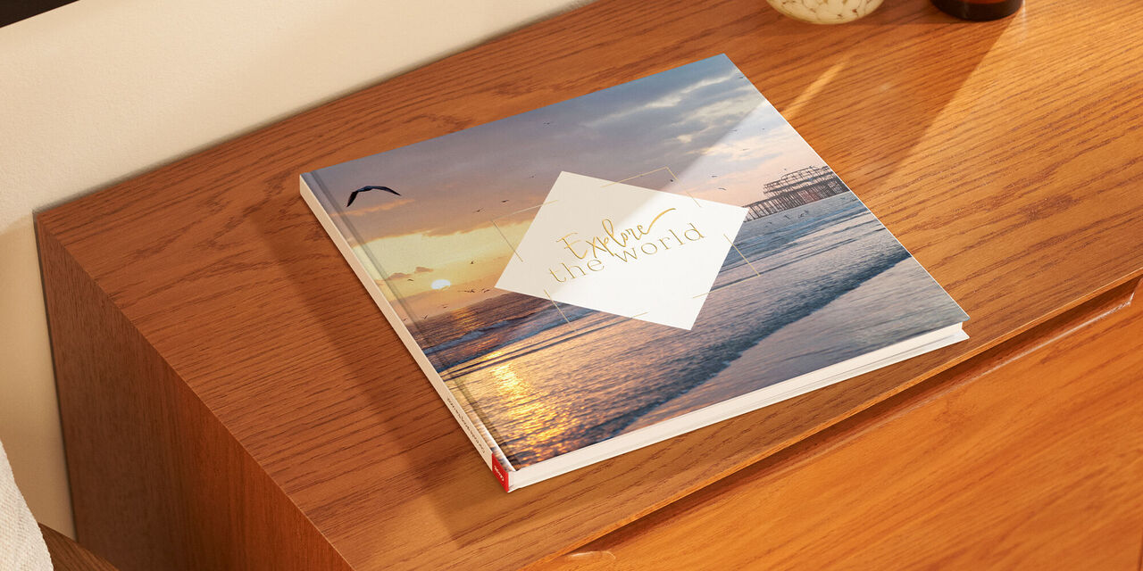 Auf einem Holzschrank liegt ein geschlossenes Fotobuch. Das Cover besteht aus einem Landschaftsporträt von einer Küste mit Pier. Im Hintergrund geht die Sonne unter. In der Mitte befindet sich ein rautenförmiges Designelement, in dem der Text „Explore the World“ steht.