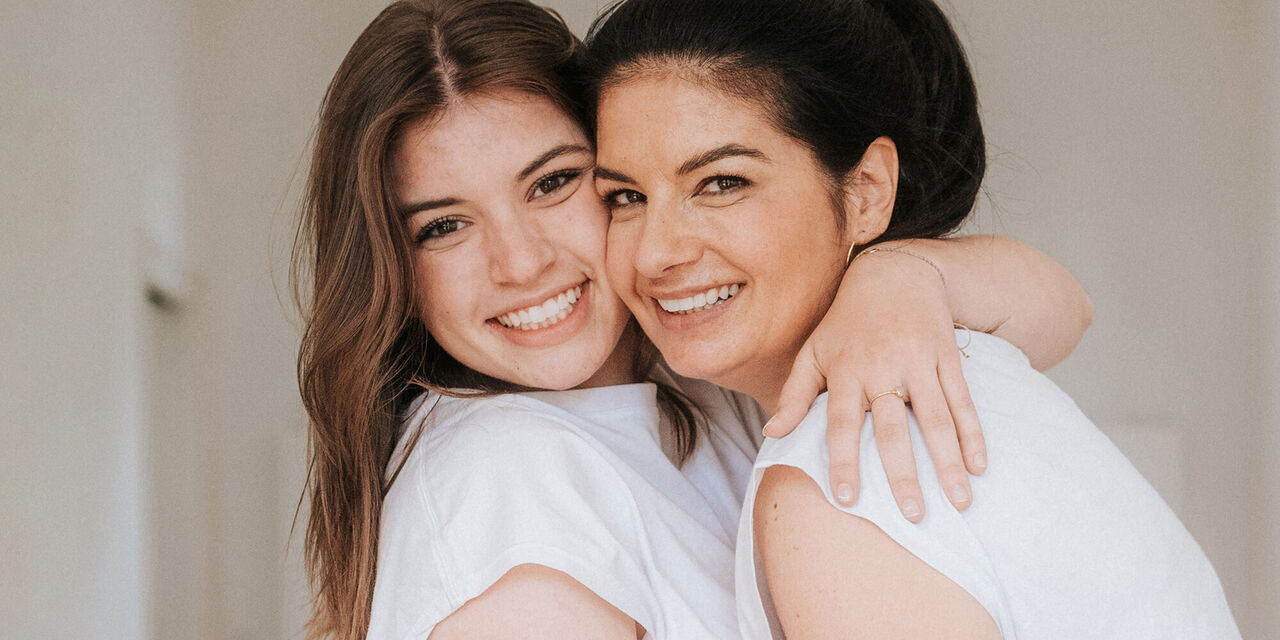 Zwei Frauen in weißen T-Shirts und grauen Jeans umarmen sich und lächeln in die Kamera.
