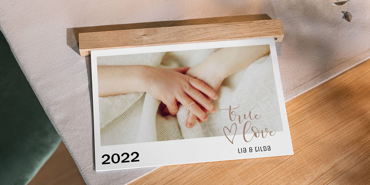 Auf einem Tisch aus Holz liegt ein Wandkalender im Querformat mit Babymotiven. Sichtbar ist das Titelbild 2022. Der Kalender wird oben von einer Holzleiste gehalten, die die Ringbindung verdeckt.