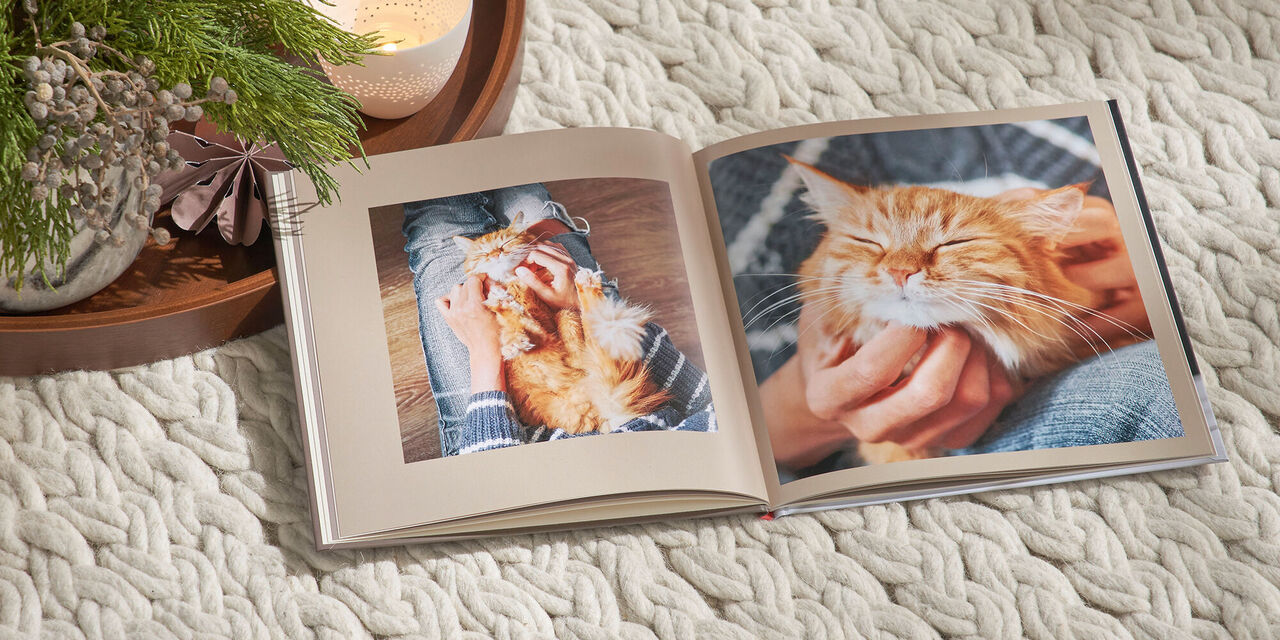 Ein aufgeschlagenes CEWE FOTOBUCH im quadratischen Format liegt auf einer weißen Decke. Auf der Doppelseite sind Fotos einer roten Katze zu sehen. Links neben dem Fotobuch liegt ein Tablett mit weihnachtlicher Deko und Kerzen.